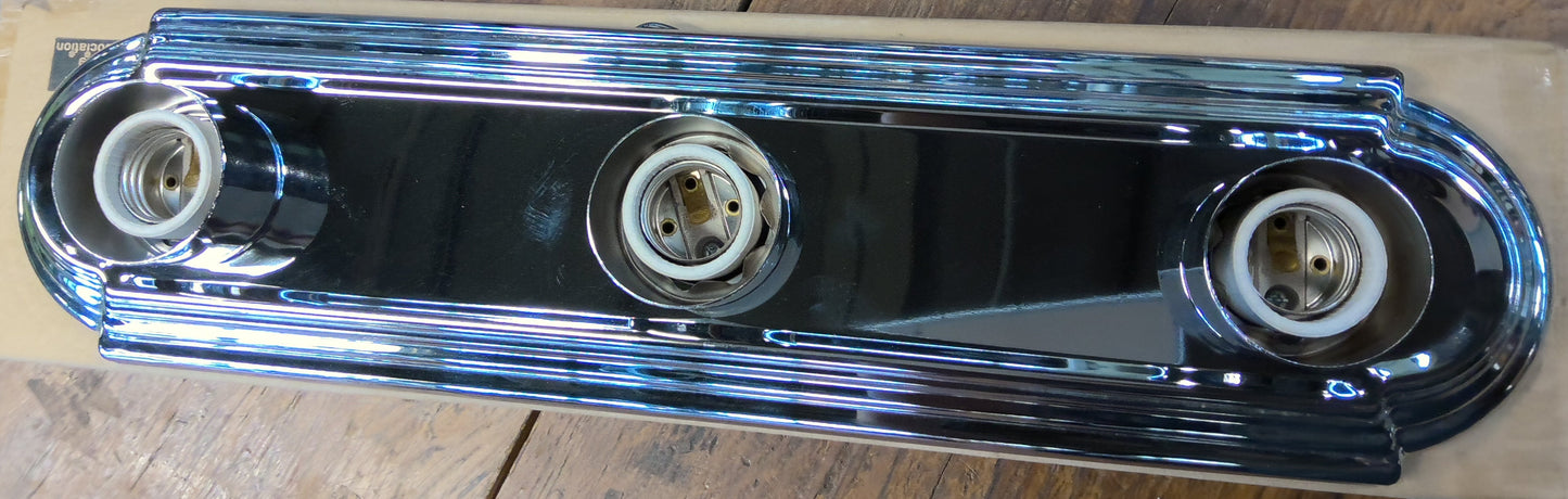 A chrome-plated metal automotive light bar with three circular bulb sockets, resting on a piece of cardboard over a rustic wooden table.