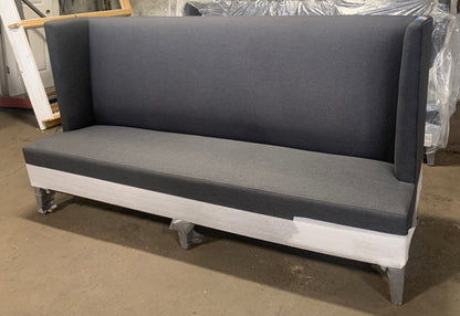 A dark gray upholstered bench with a tall backrest and light gray base, photographed in a workshop or warehouse setting with other furniture pieces visible in the background.