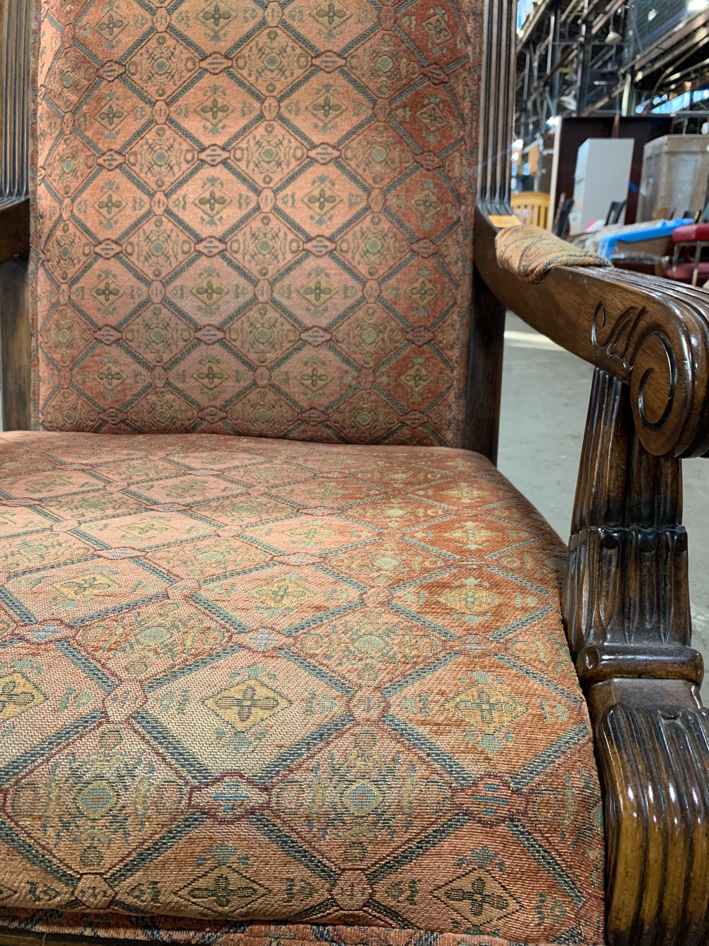 Close-up of an upholstered wooden armchair with a patterned fabric seat and backrest in warm beige, rust, and muted green tones, set in what appears to be a warehouse or furniture showroom.