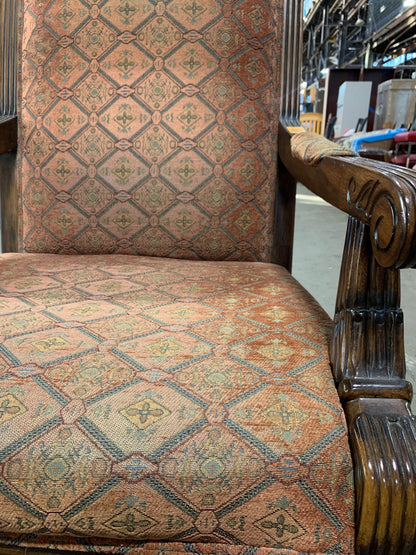 Close-up of an upholstered wooden armchair with a patterned fabric seat and backrest in warm beige, rust, and muted green tones, set in what appears to be a warehouse or furniture showroom.