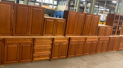 A row of medium brown wooden kitchen cabinets, including upper and lower cabinets with paneled doors and drawer fronts, displayed in a warehouse setting.