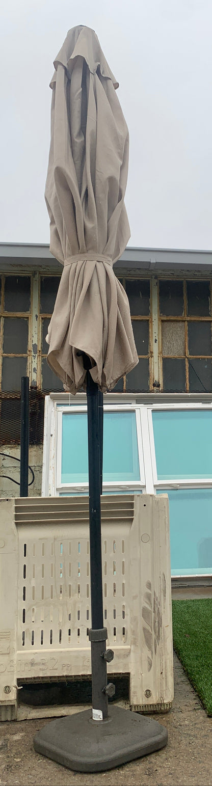 A closed beige patio umbrella with a black pole stands next to a cream-colored outdoor lounge chair on a patio in front of a building with large windows.