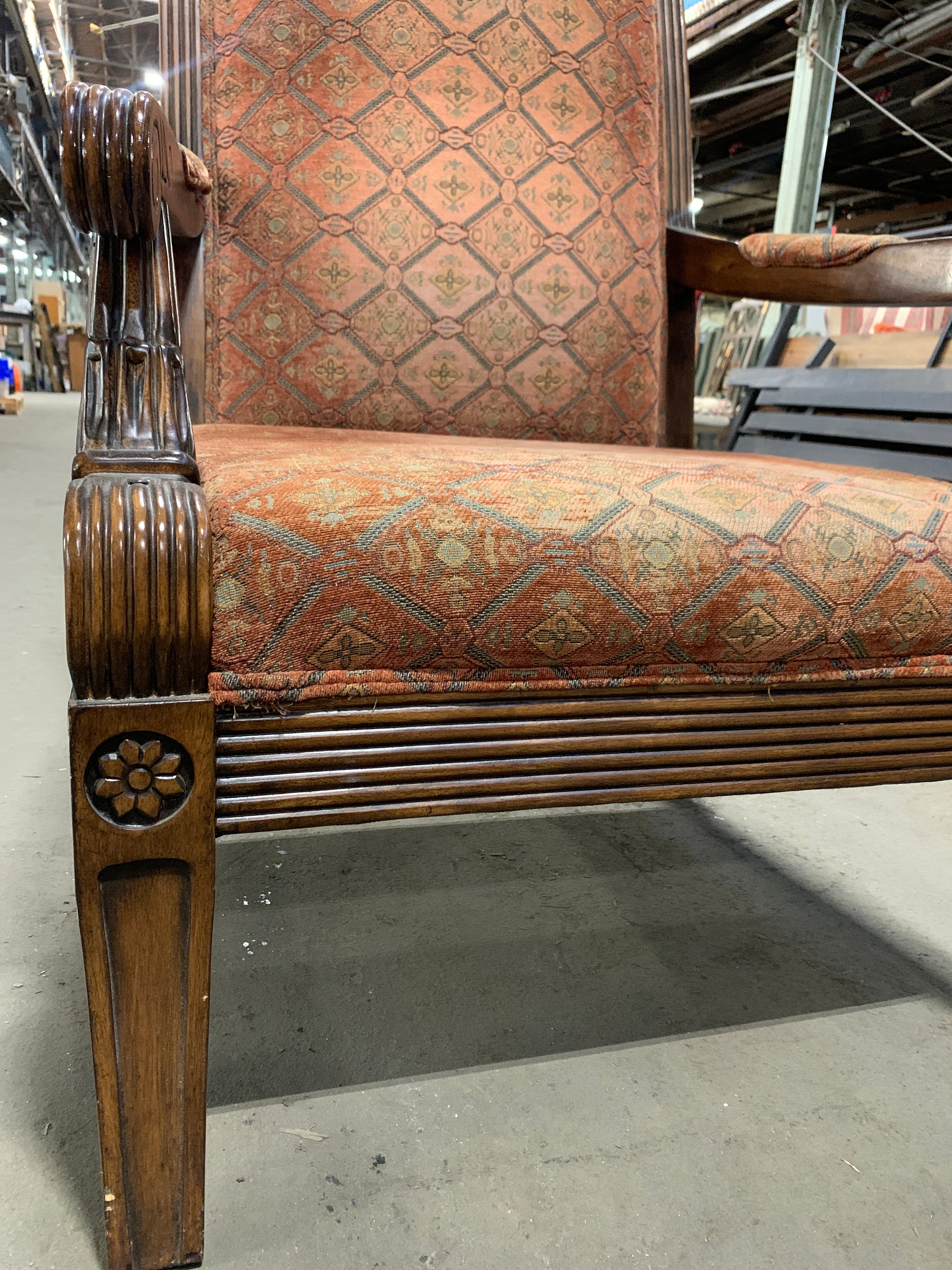 A close-up of an antique wooden armchair with carved details, featuring rust-orange patterned fabric upholstery on the seat and backrest, photographed in a warehouse setting.