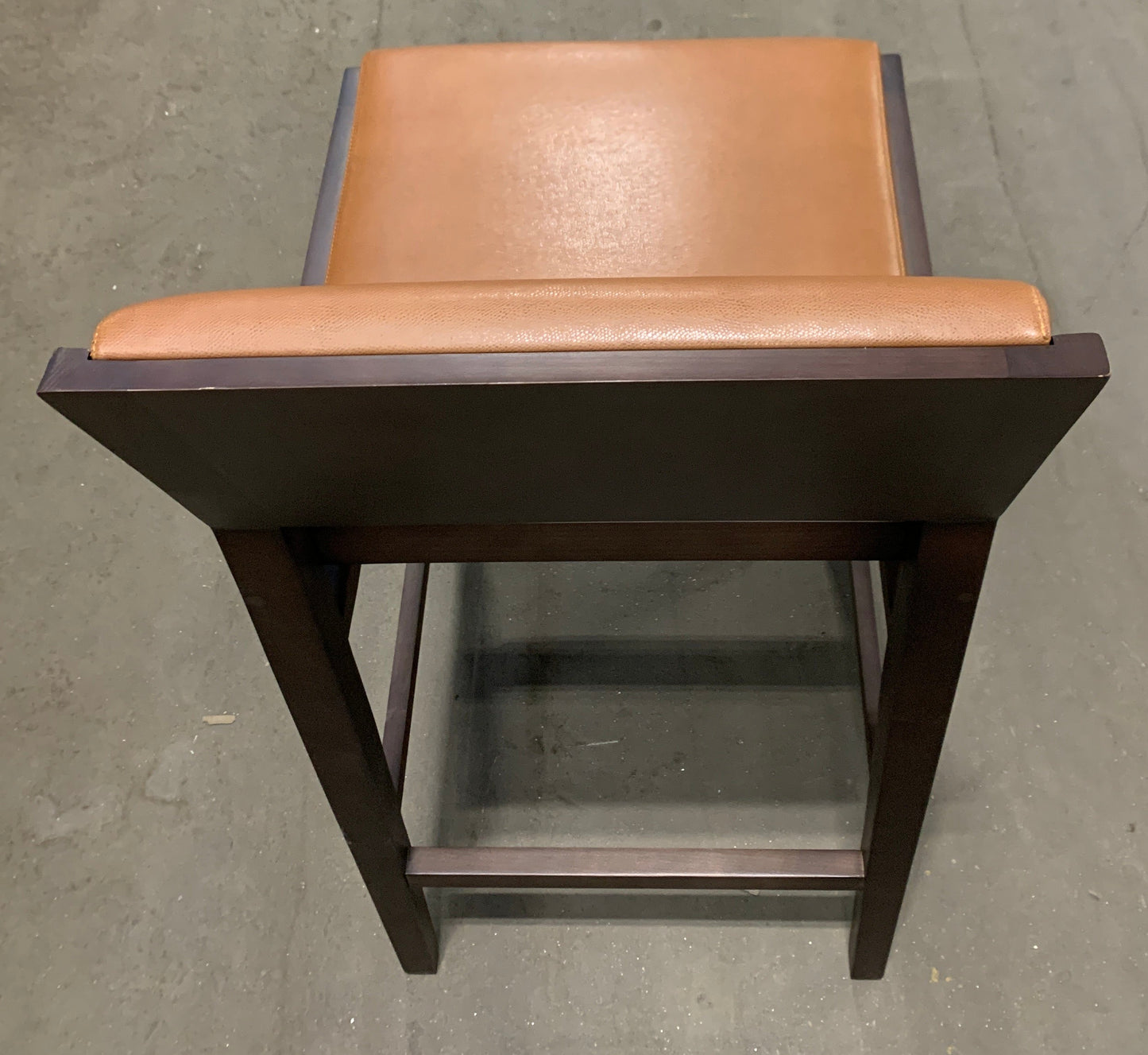 A bar stool with a dark brown wooden frame and a tan leather seat cushion, photographed from a top-down angle on a concrete floor.