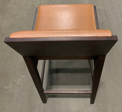 A bar stool with a dark brown wooden frame and a tan leather seat cushion, photographed from a top-down angle on a concrete floor.