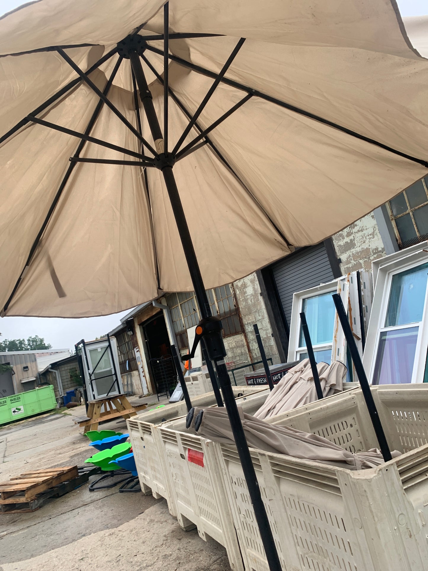 A large beige patio umbrella with a black metal frame and pole, photographed in an outdoor warehouse or showroom setting with other umbrellas and storage items visible in the background.
