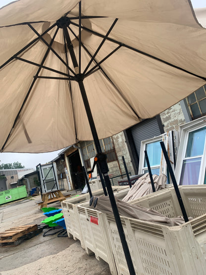 A large beige patio umbrella with a black metal frame and pole, photographed in an outdoor warehouse or showroom setting with other umbrellas and storage items visible in the background.
