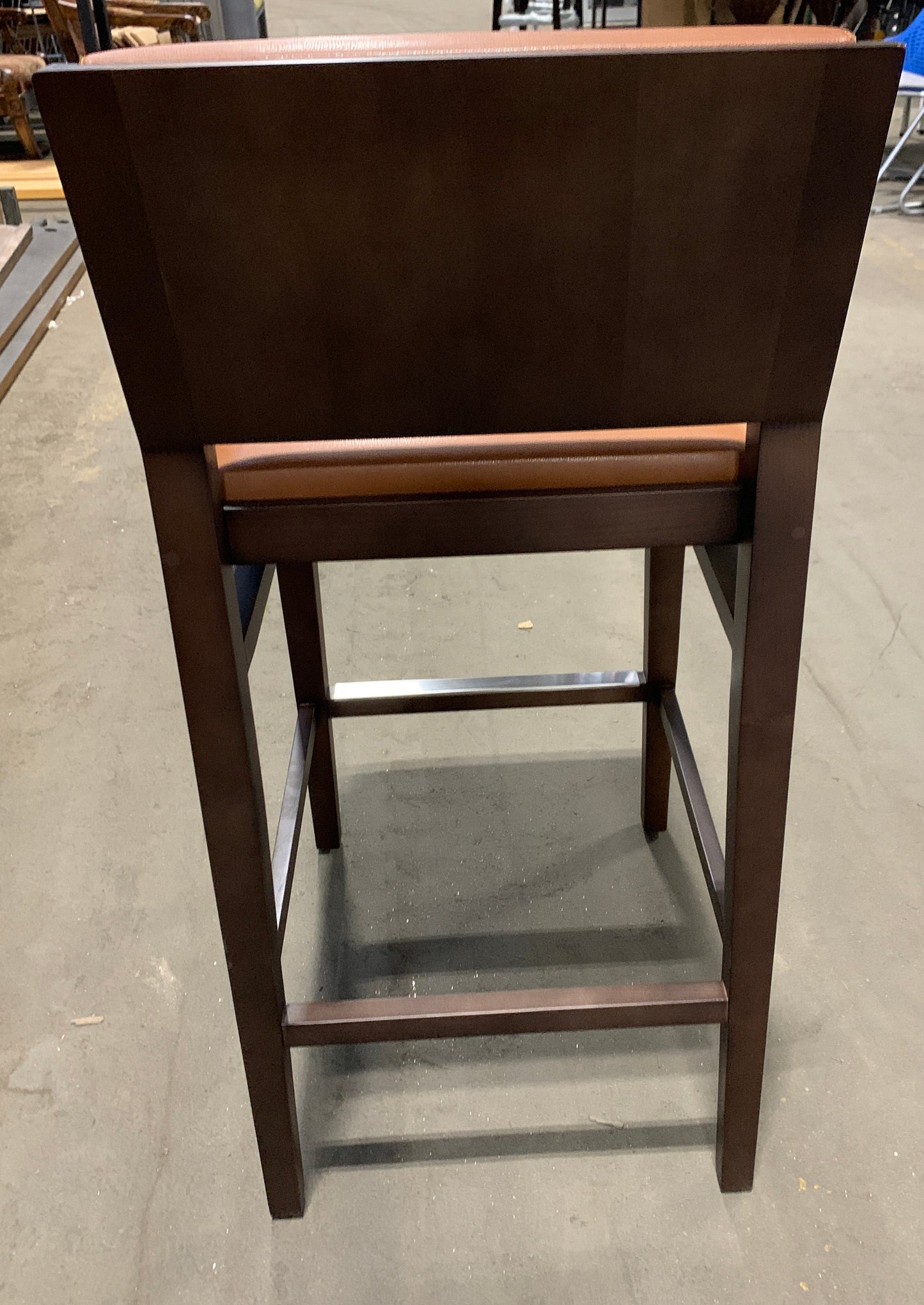 A dark brown wooden barstool with a high back and a brown upholstered seat cushion, photographed from the rear in what appears to be a furniture showroom or warehouse.