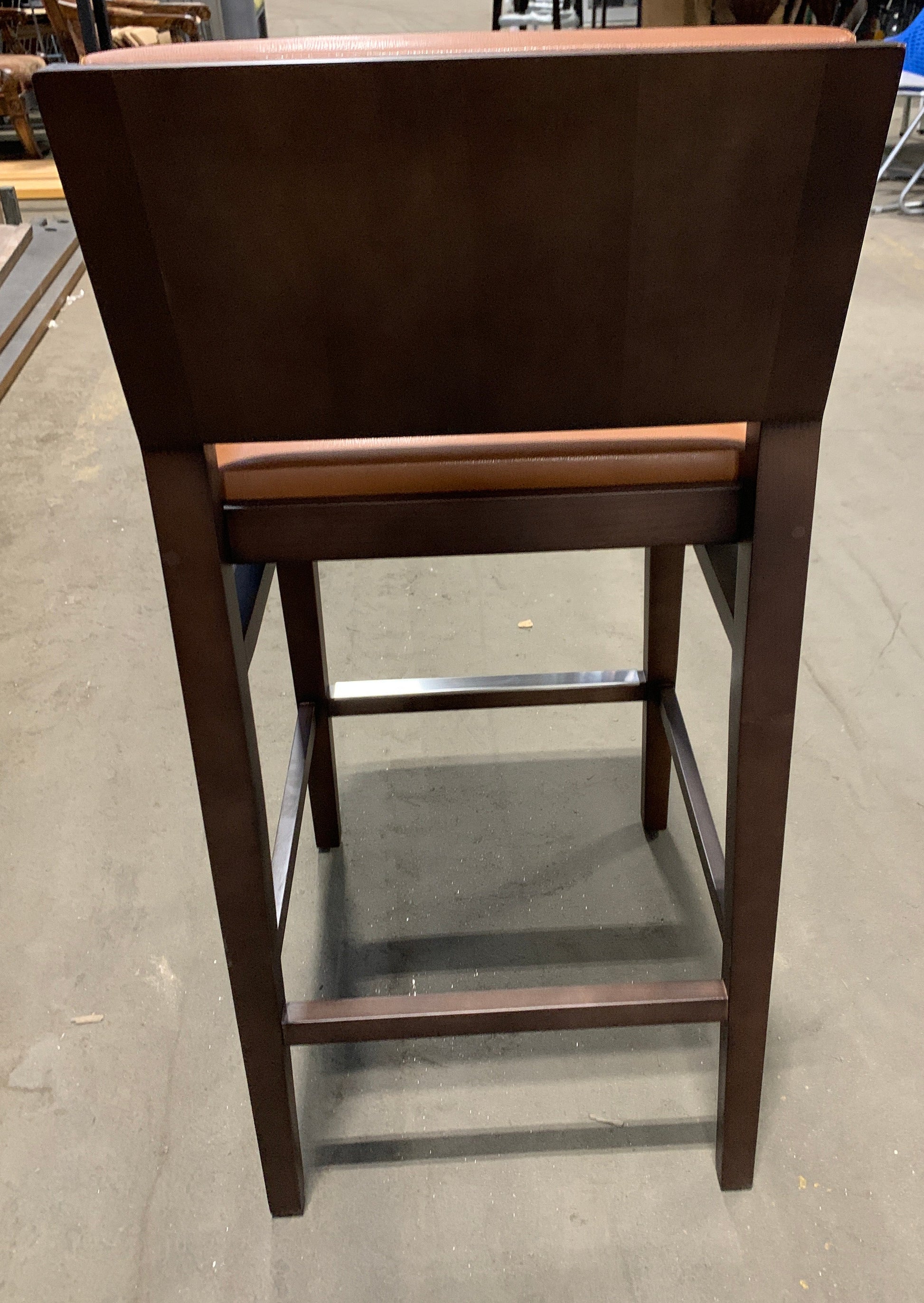 A dark brown wooden barstool with a high back and a brown upholstered seat cushion, photographed from the rear in what appears to be a furniture showroom or warehouse.