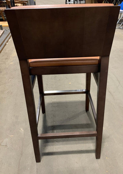 A dark brown wooden barstool with a high back and a brown upholstered seat cushion, photographed from the rear in what appears to be a furniture showroom or warehouse.