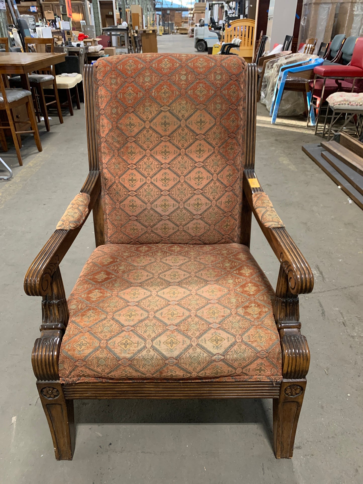 An antique-style wooden armchair with carved dark brown wood arms and legs, upholstered in a rust-orange and beige diamond-patterned fabric, photographed in a furniture store.