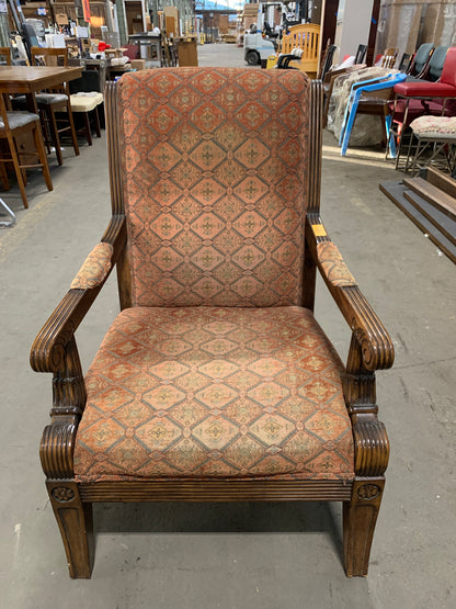 An antique-style wooden armchair with carved dark brown wood arms and legs, upholstered in a rust-orange and beige diamond-patterned fabric, photographed in a furniture store.