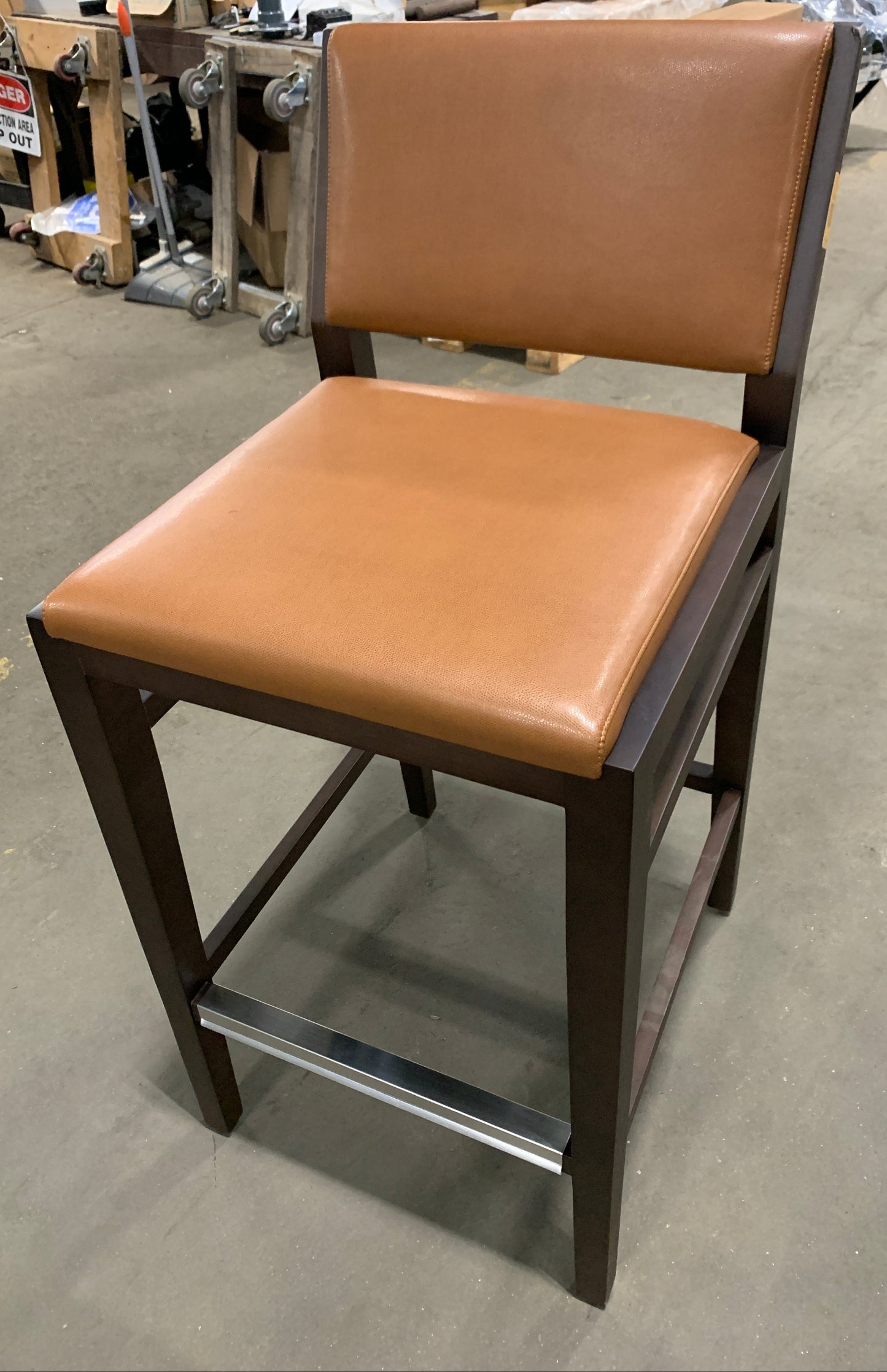 A wooden bar stool with a tan leather seat and backrest, featuring a dark brown wood frame and a chrome metal footrest, photographed in a warehouse setting.