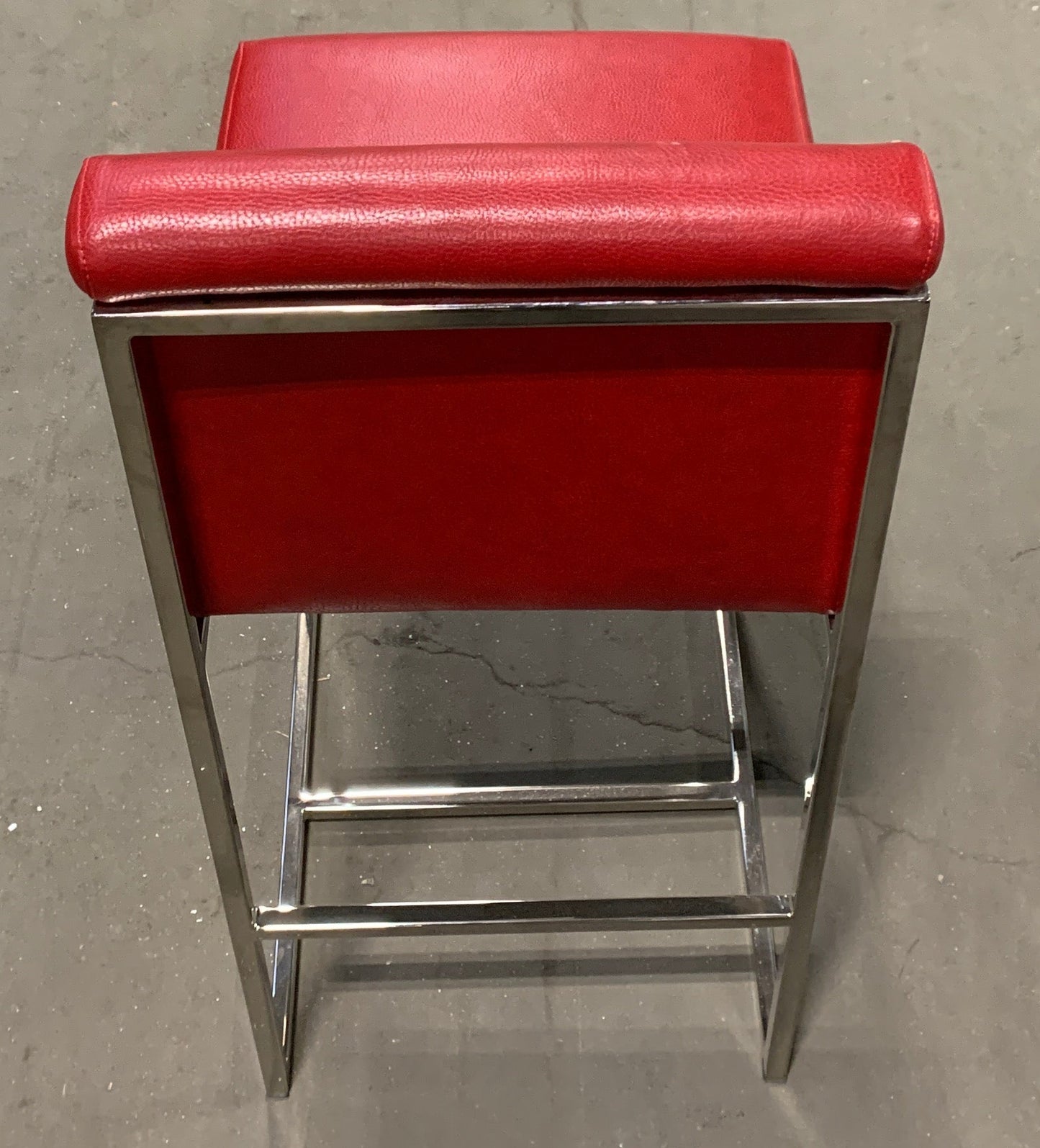 A red upholstered bar stool with a chrome metal frame, photographed from behind in what appears to be a warehouse or furniture store.