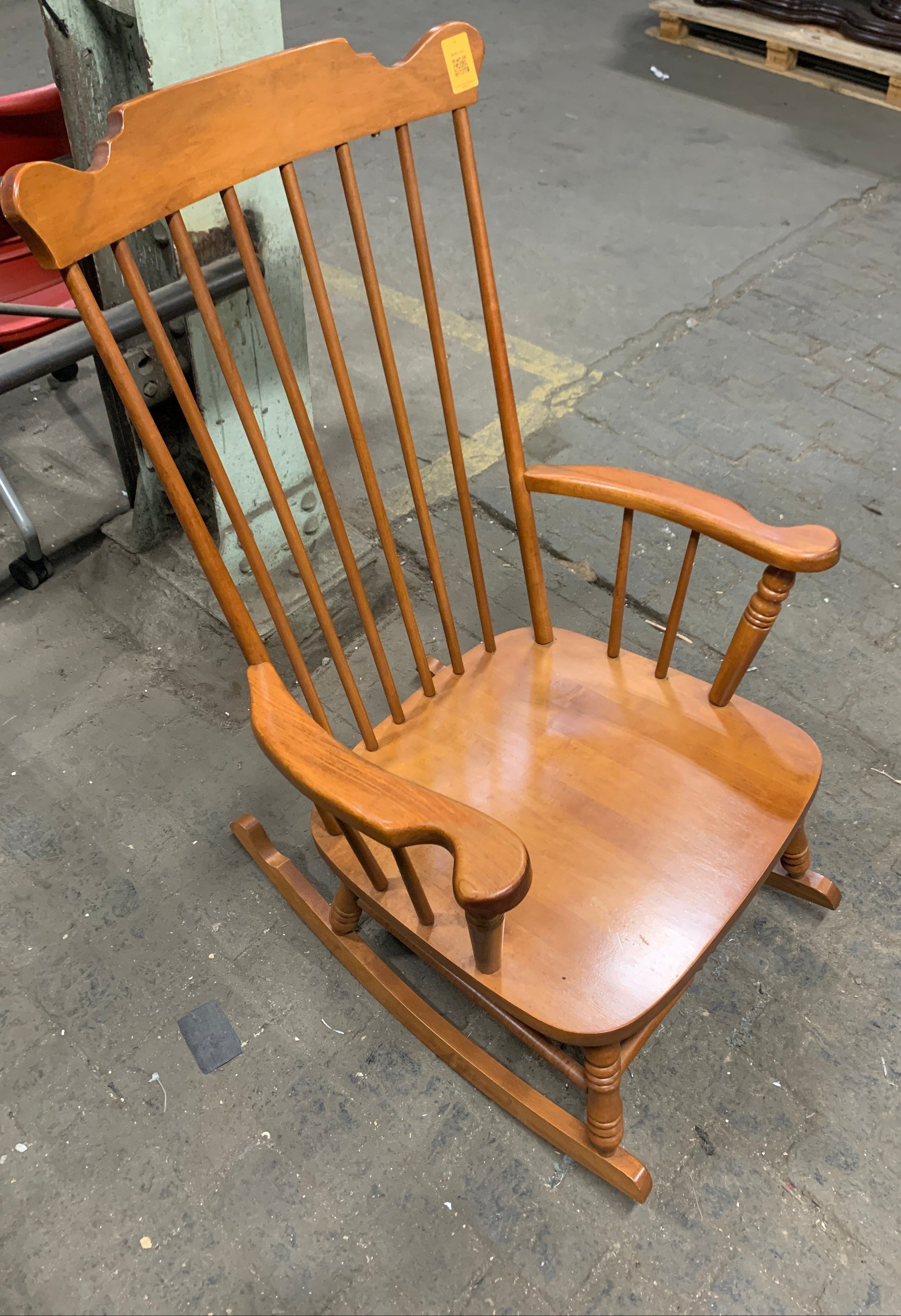 A wooden rocking chair with a spindle-back design, photographed at an angle on a concrete floor.