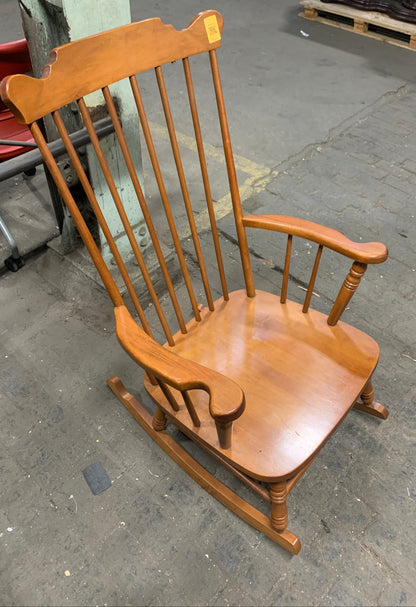 A wooden rocking chair with a spindle-back design, photographed at an angle on a concrete floor.