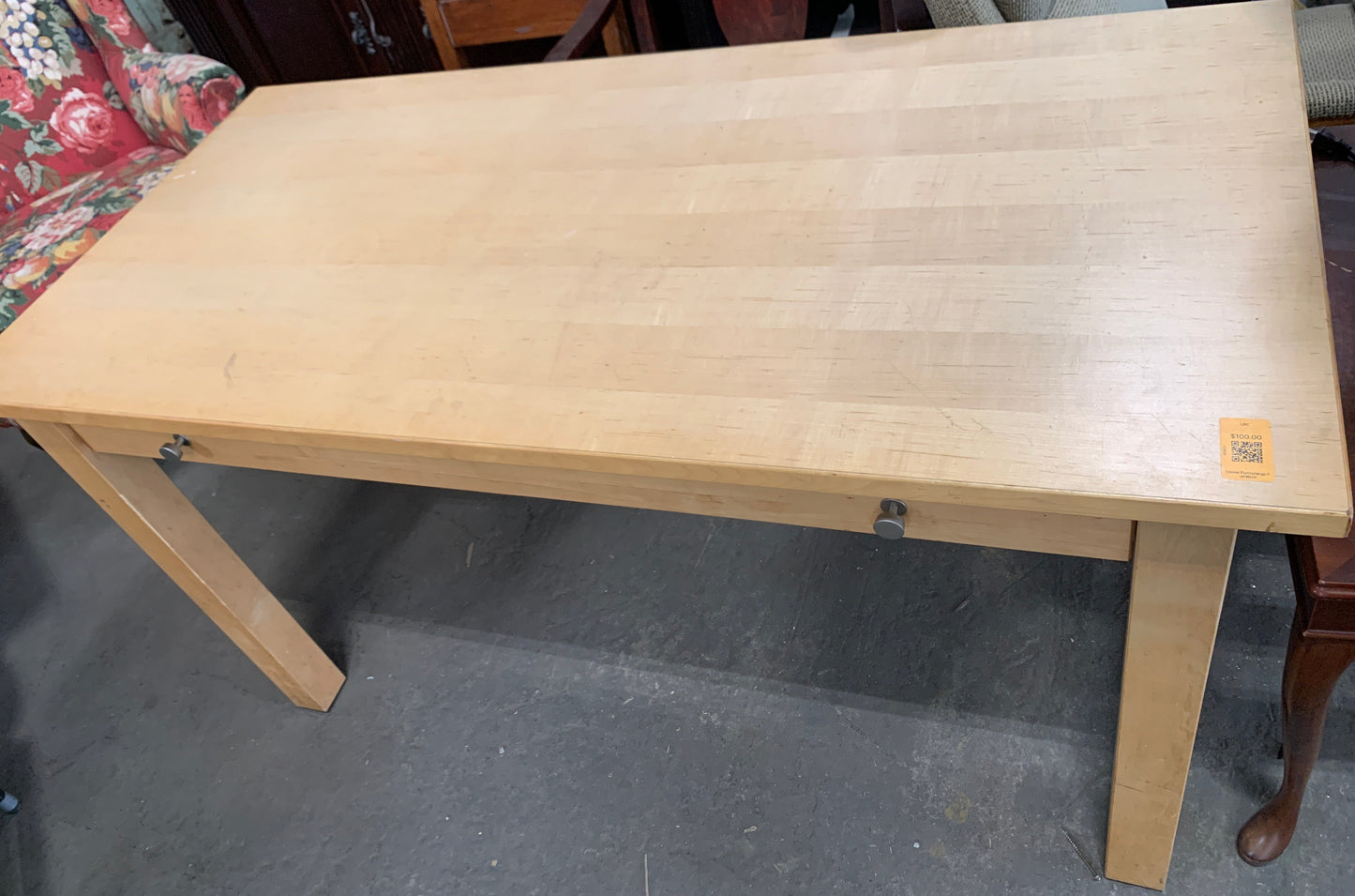 A light natural wood dining table with a rectangular top and four legs, photographed in what appears to be a furniture store or warehouse setting.