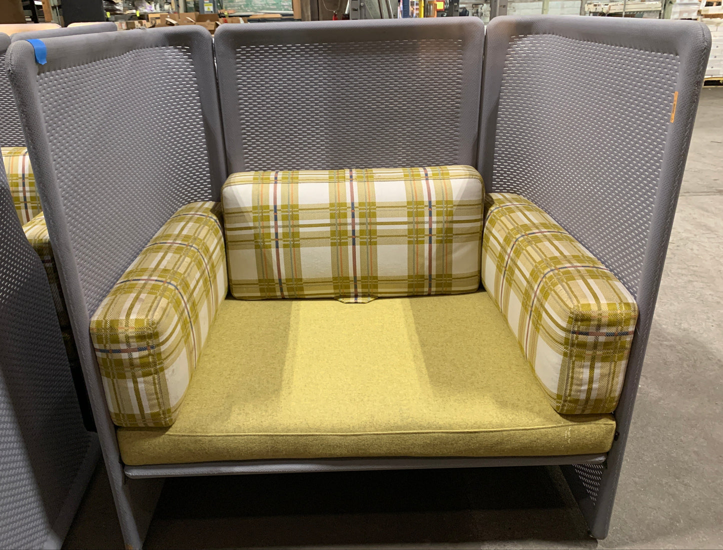 A modern gray mesh high-back lounge chair with yellow-green plaid armrests and a matching yellow-green seat cushion, photographed in a warehouse showroom.