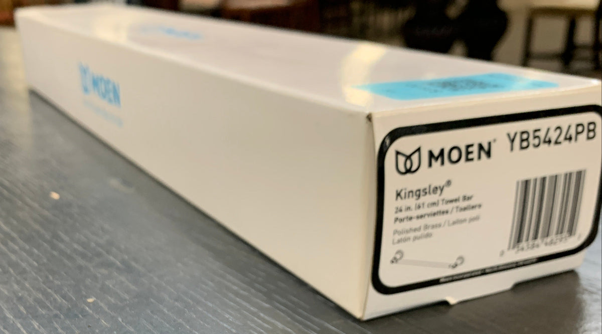 A white Moen product box with a barcode label, photographed on a textured gray tabletop in a real indoor setting with blurred background elements.