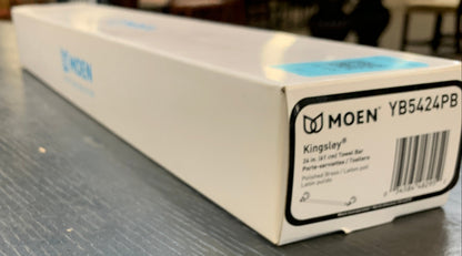 A white Moen product box with a barcode label, photographed on a textured gray tabletop in a real indoor setting with blurred background elements.