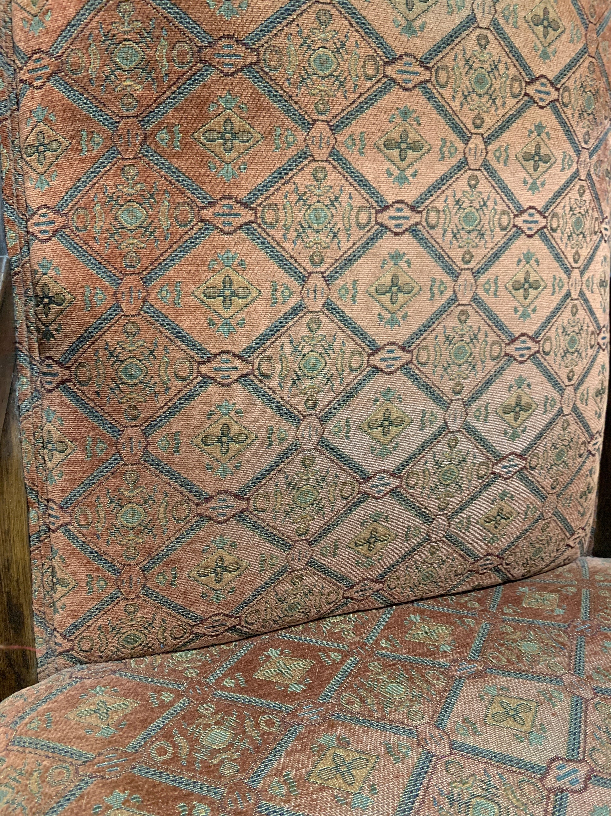 Close-up of patterned upholstery fabric on a chair or upholstered furniture piece, showing a repeating diamond motif in muted teal, rust, beige, and brown tones.