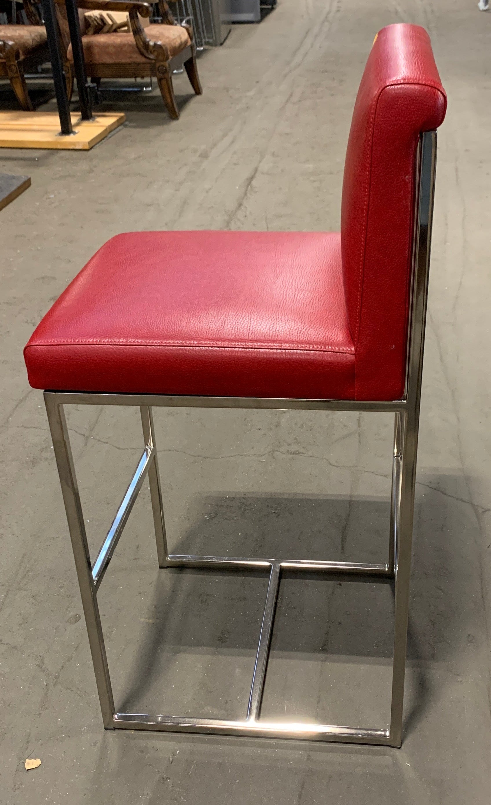 A modern bar stool with a red upholstered seat and backrest on a polished chrome metal frame, photographed in a warehouse or furniture store setting.