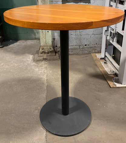 A round wooden pub table with a warm orange-brown wood top and a black metal pedestal base, photographed in a warehouse or showroom setting.