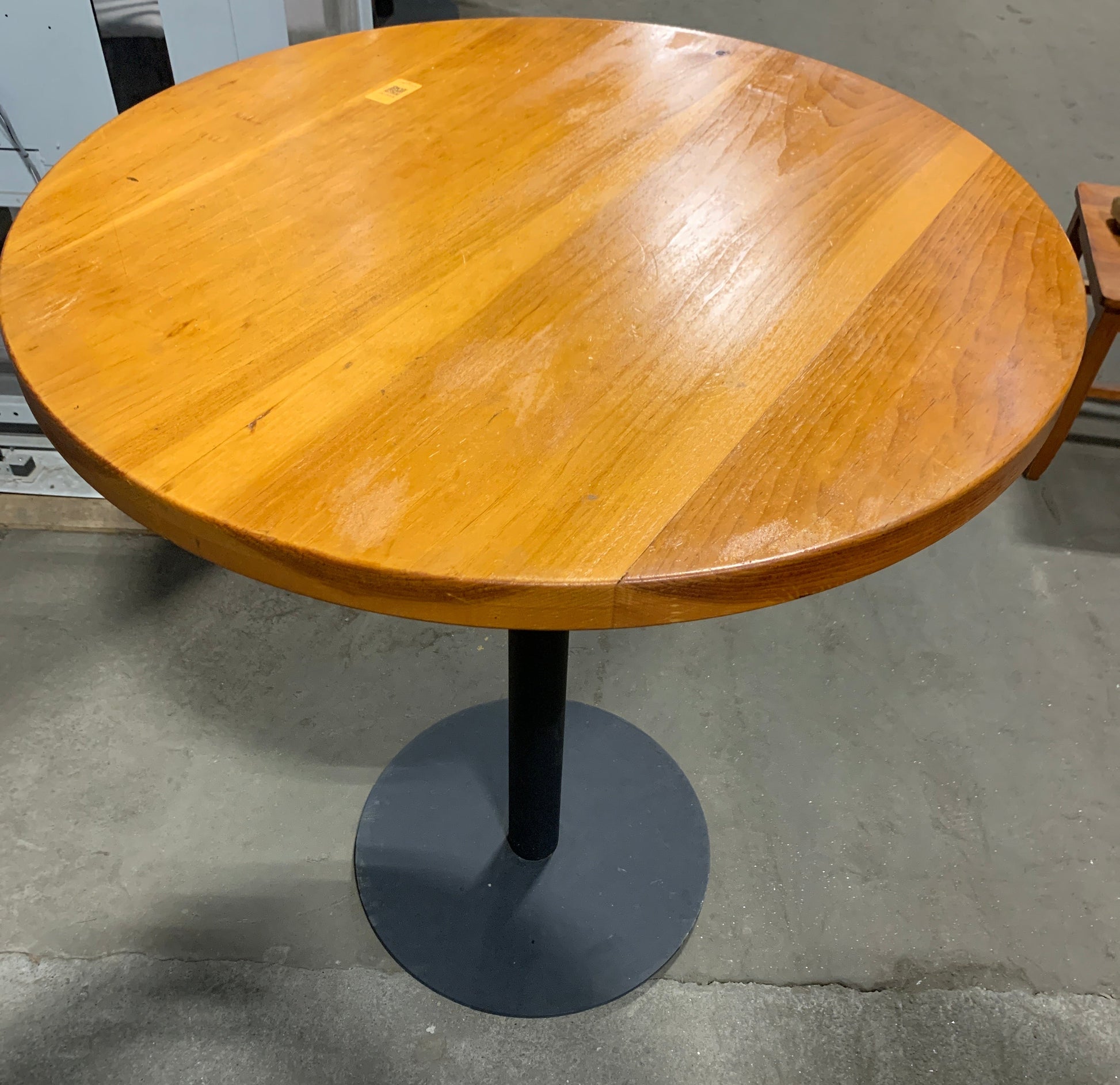 Round wooden pub table with a warm honey finish and a black metal pedestal base, photographed in a warehouse setting.