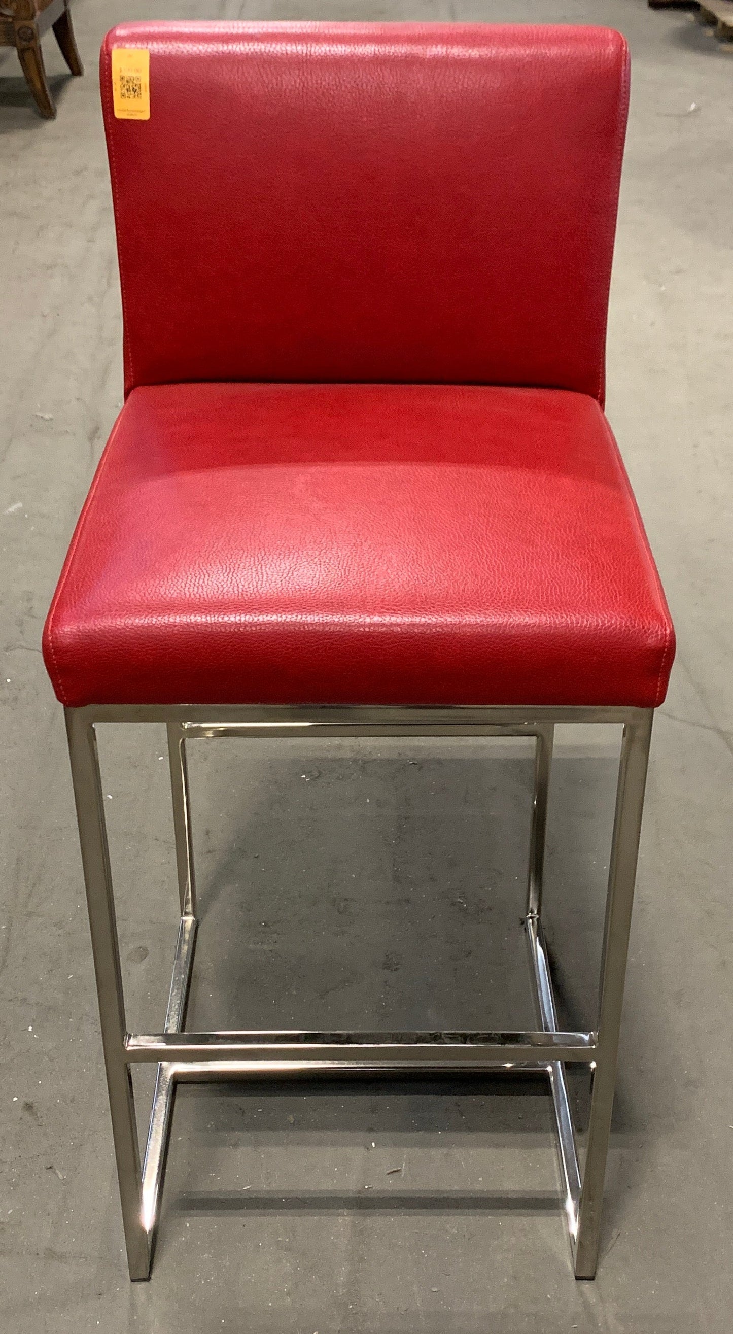 A red upholstered bar stool with a chrome metal frame, photographed in a furniture store aisle.