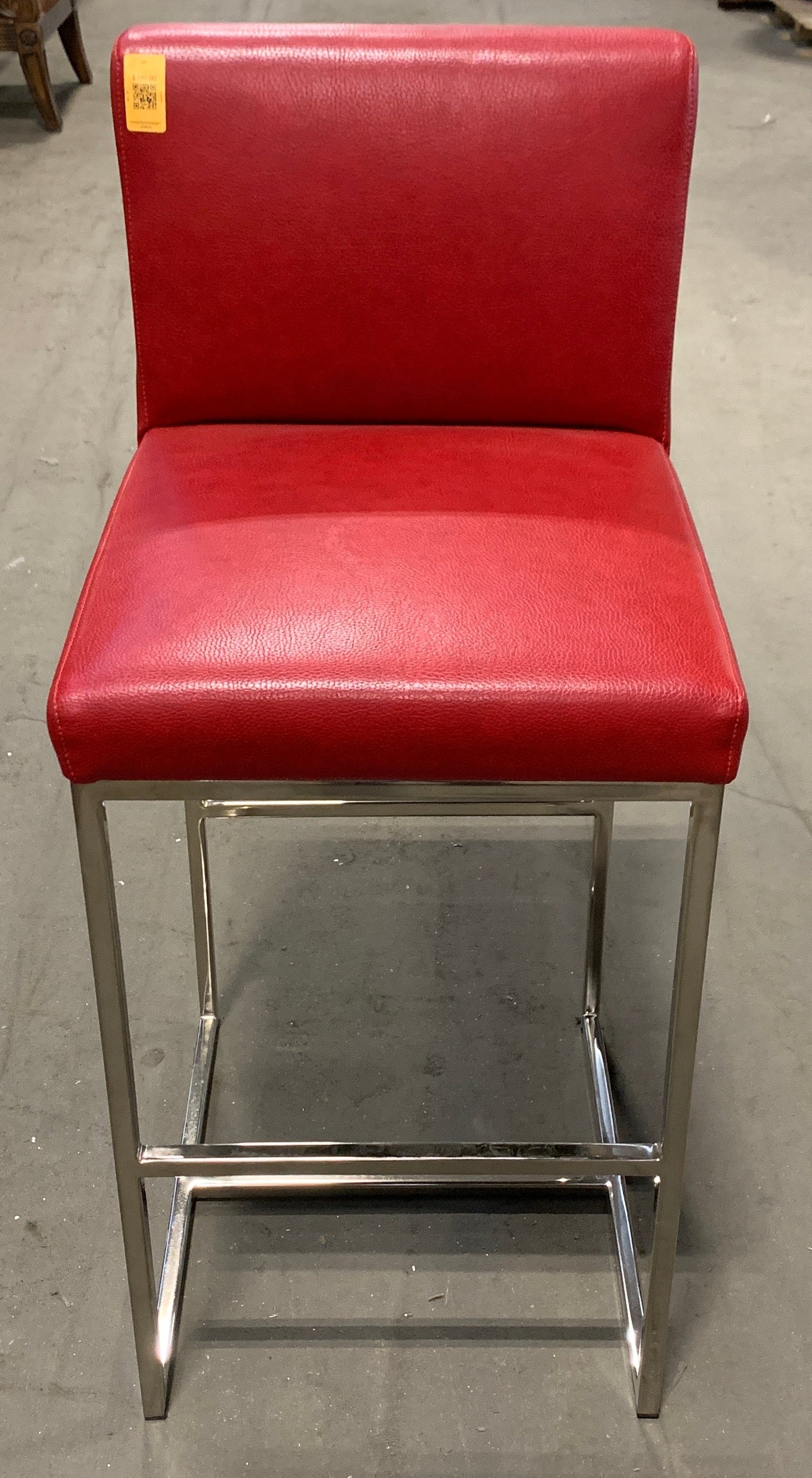 A red upholstered bar stool with a chrome metal frame, photographed in a furniture store aisle.