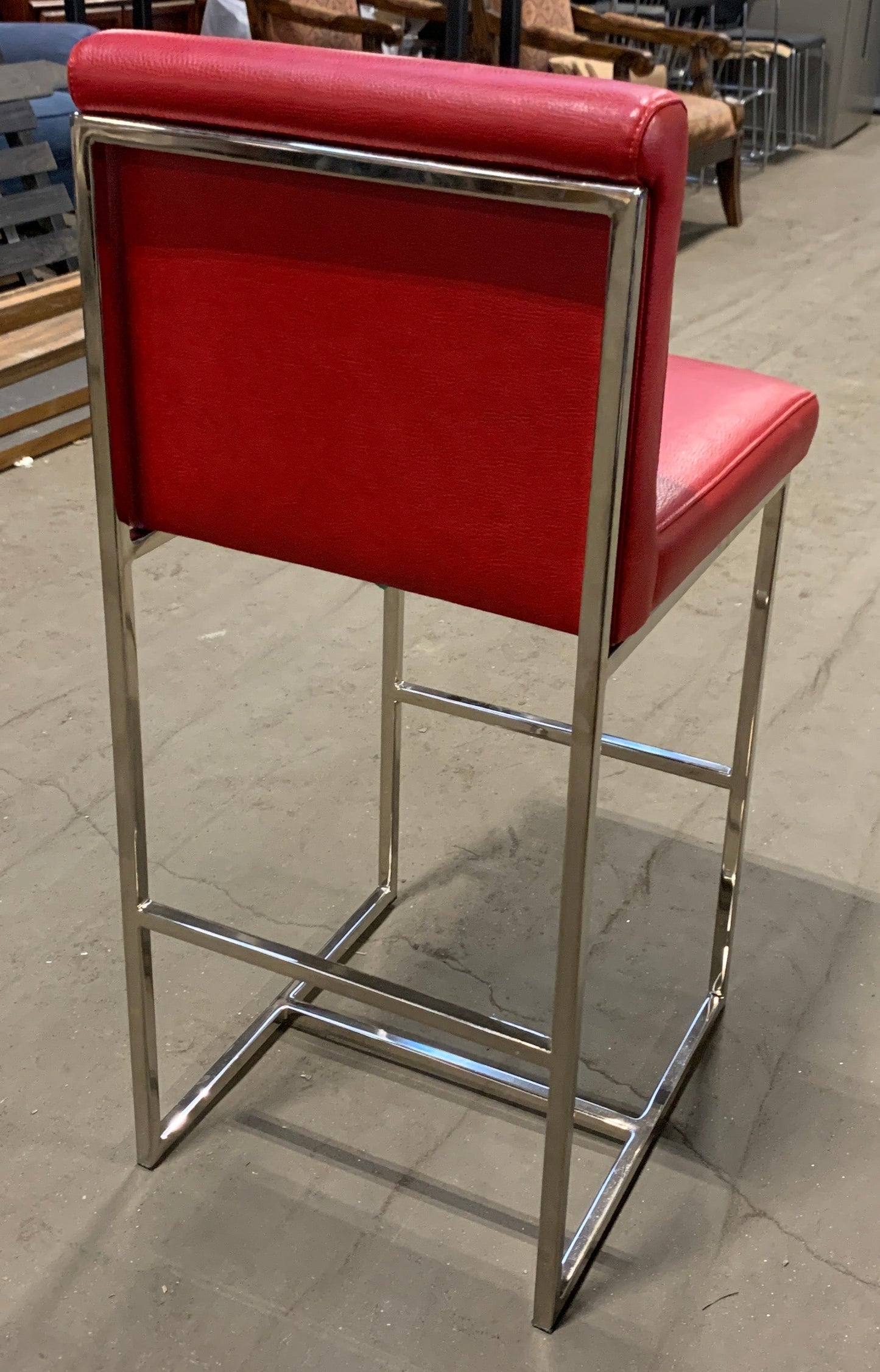 A modern bar stool with a bright red upholstered seat and backrest, supported by a polished chrome metal frame, photographed in a furniture showroom.