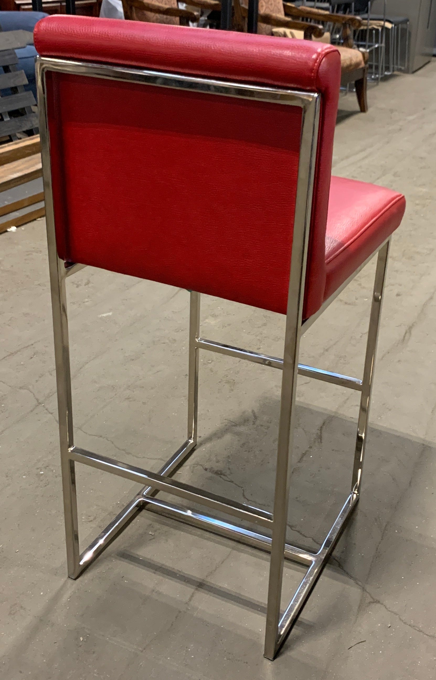 A modern bar stool with a bright red upholstered seat and backrest, supported by a polished chrome metal frame, photographed in a furniture showroom.