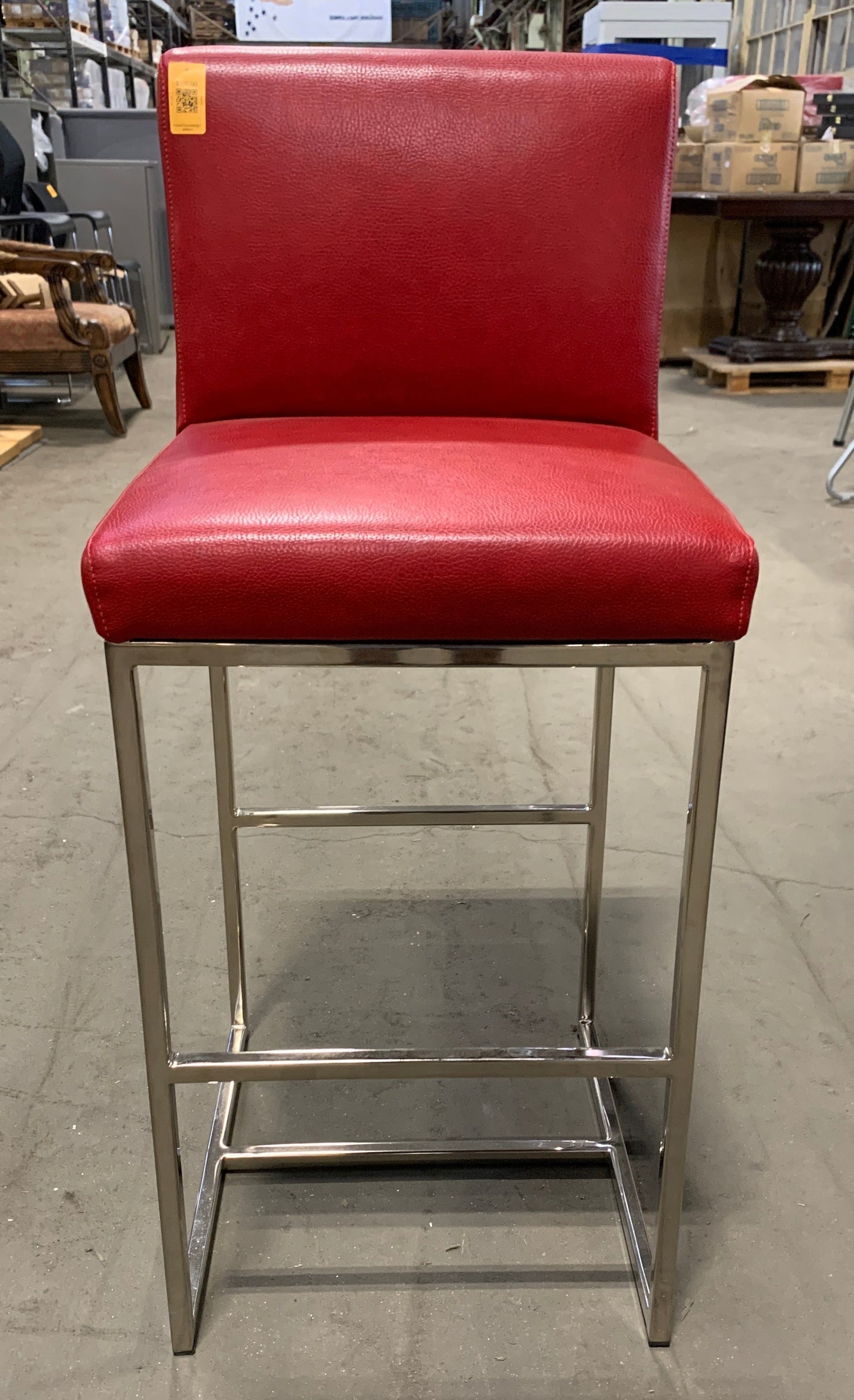 A red upholstered bar stool with a square backrest and a chrome metal frame, photographed in a furniture store showroom.
