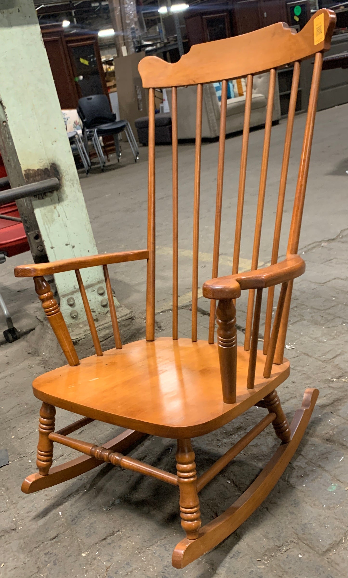 A wooden spindleback rocking chair with a warm medium-brown finish, photographed in what appears to be a warehouse or furniture showroom on a concrete floor.