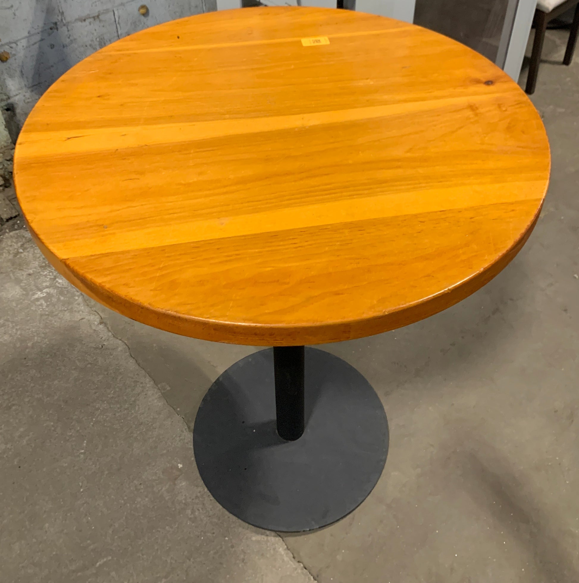 A round wooden bar table with a warm honey-orange finish and a black metal pedestal base, photographed in a warehouse or showroom setting.