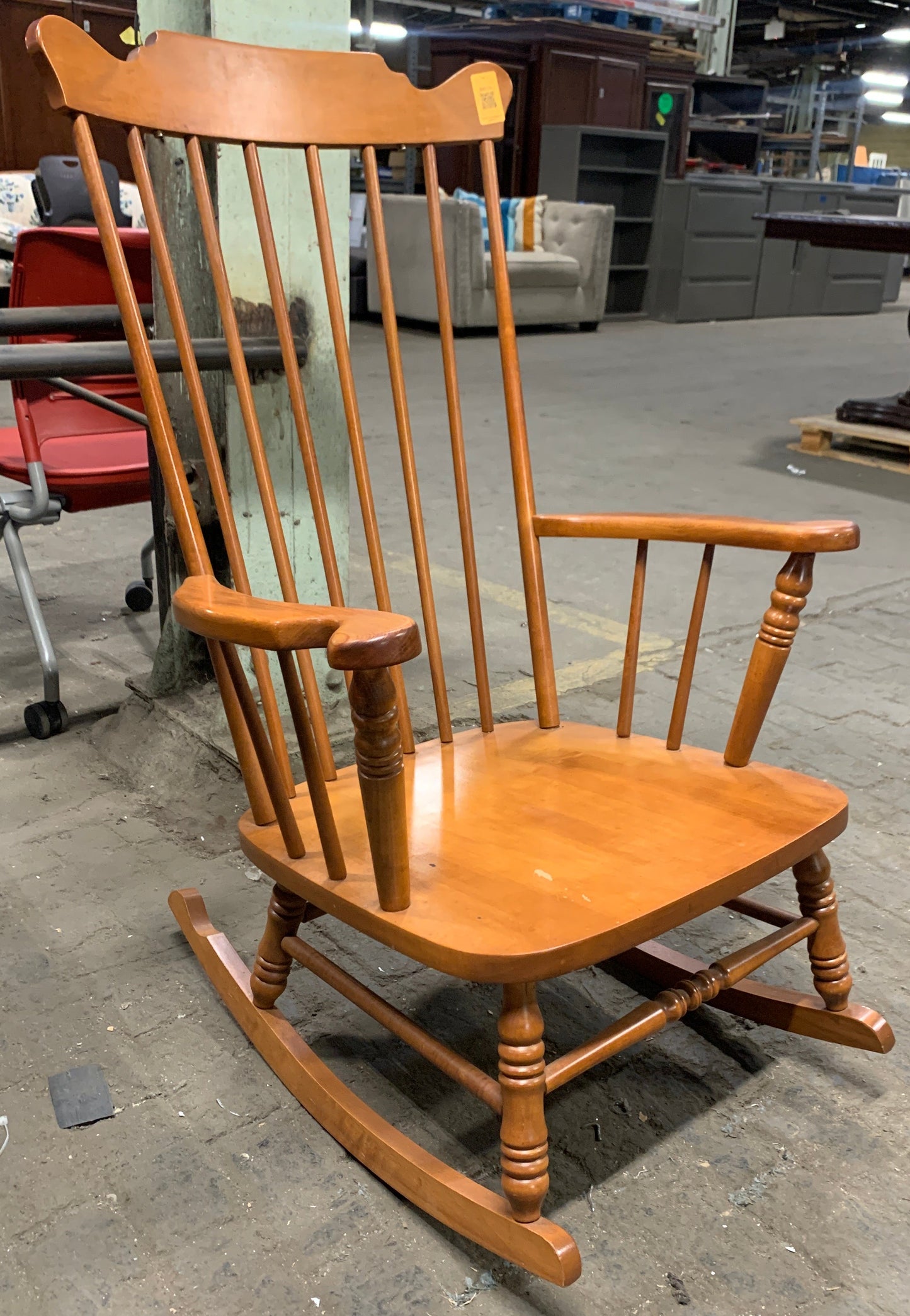 A wooden rocking chair with a spindle-back design, photographed in what appears to be a warehouse or furniture showroom on a concrete floor.