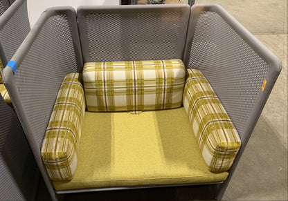 A gray mesh high-back lounge chair with mustard yellow and white plaid armrests and a matching yellow seat cushion, photographed in a warehouse setting.