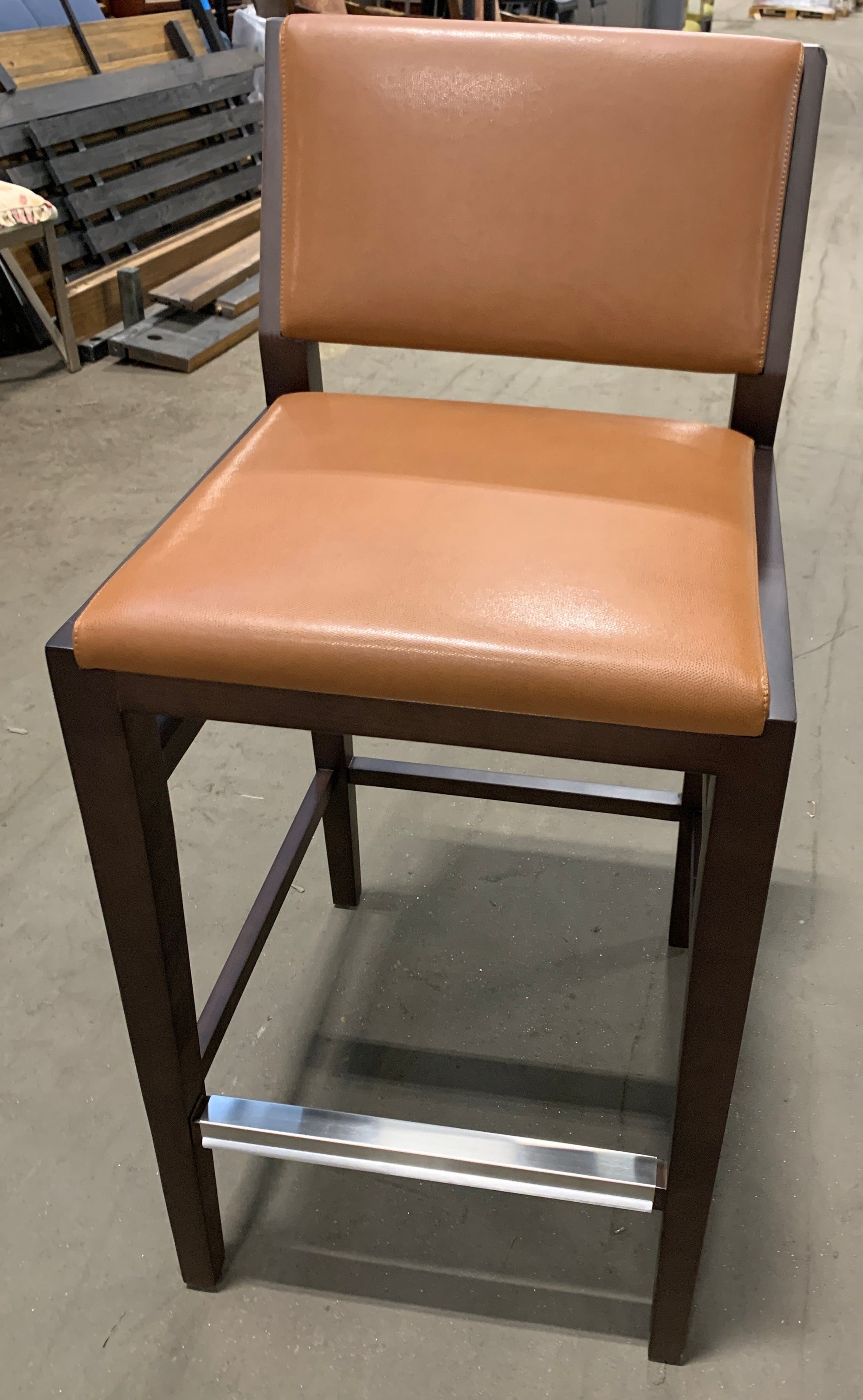 A modern bar stool with a tan leather seat and backrest, dark brown wooden frame, and a chrome footrest bar, photographed in a furniture store.