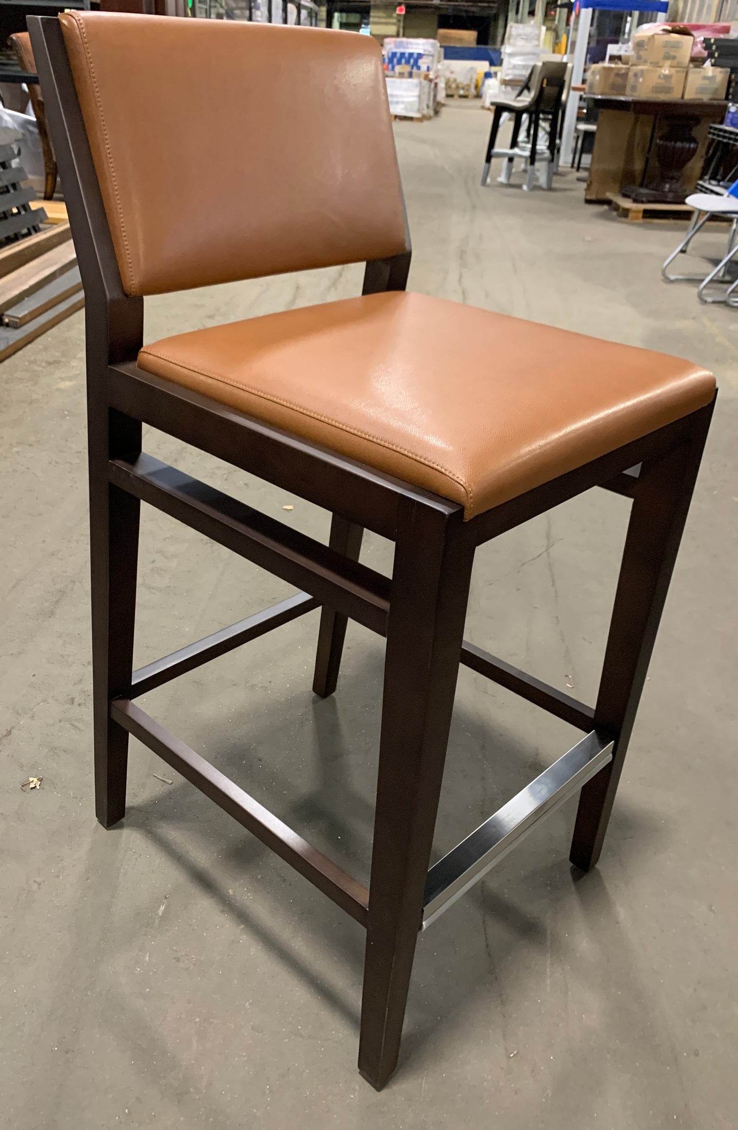 A wooden bar stool with a warm tan leather seat and backrest, set in a warehouse or furniture store environment.