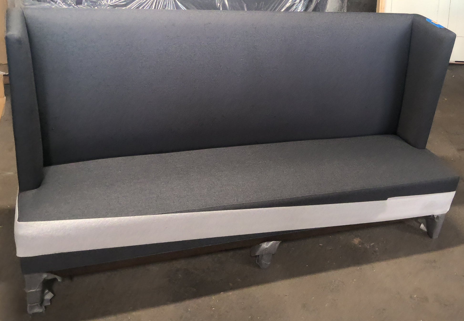 A dark gray upholstered bench or sofa with a high back and wide armrests, shown in a warehouse or workshop setting with cardboard boxes, wrapped materials, and an orange traffic cone in the background.