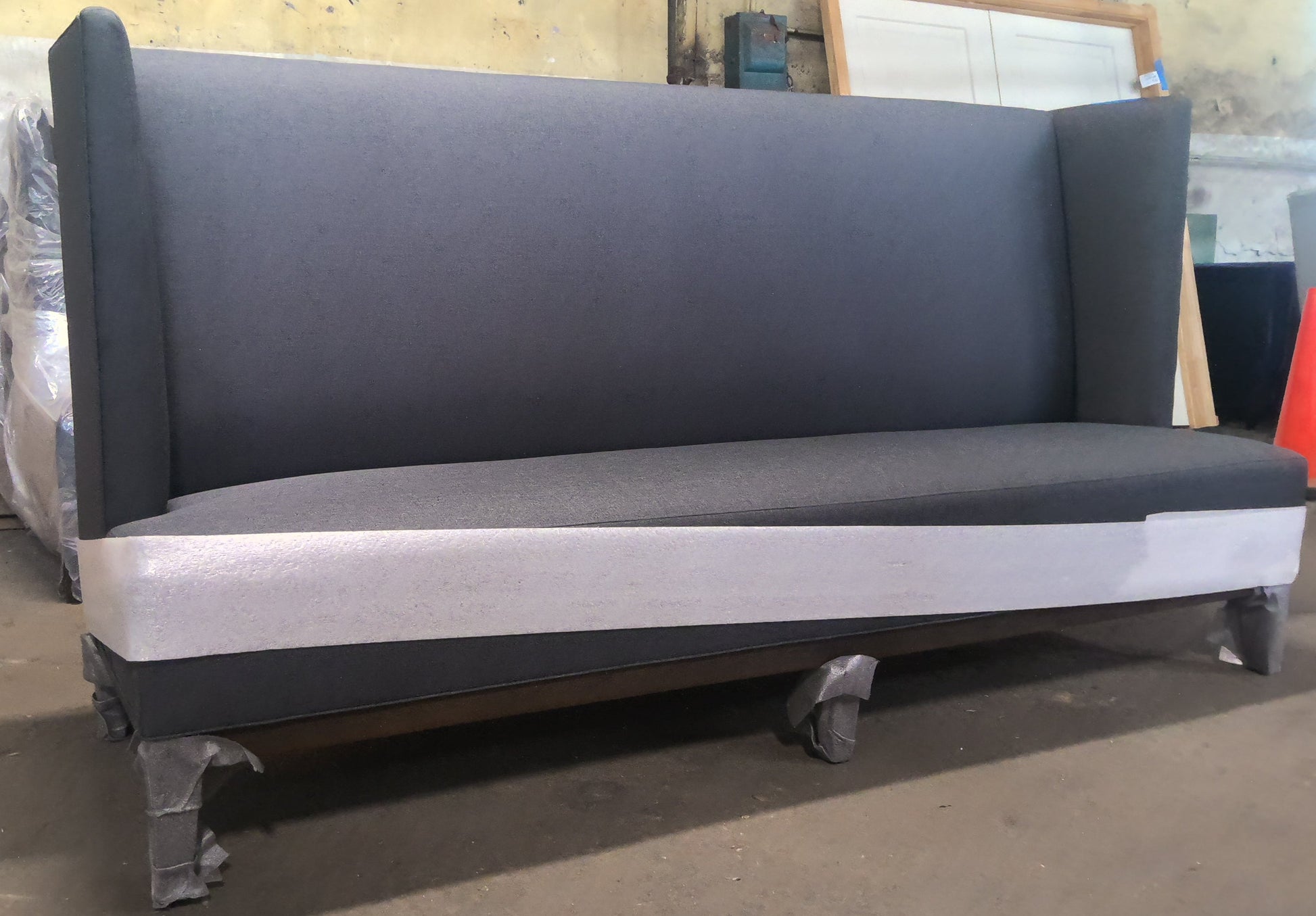 A dark gray upholstered bench or sofa with a high back and metal legs, photographed in a warehouse or workshop setting with boxes and other furniture visible in the background.