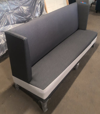 A dark gray upholstered bench or sofa with a white base, photographed in a warehouse or workshop setting with other furniture and materials visible in the background.