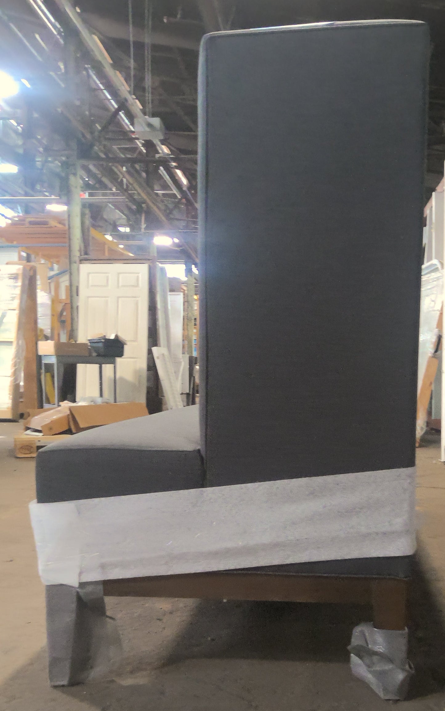 A tall upholstered dining chair with a dark gray fabric and a white protective paper wrap around the base, photographed in a warehouse or furniture workshop setting.