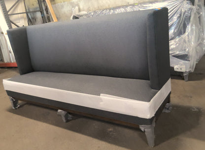 A dark gray upholstered bench with a tall backrest and white side trim, photographed in a warehouse or furniture storage area with other wrapped items in the background.