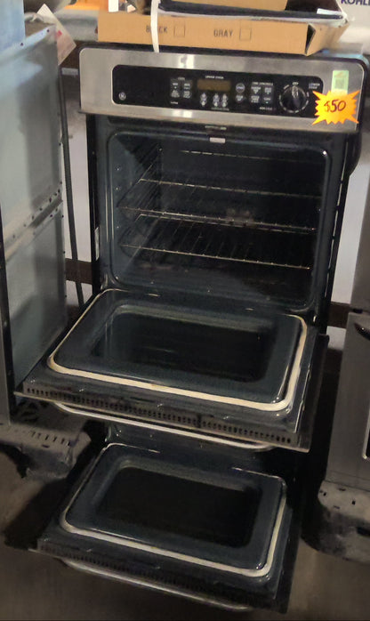 A stainless steel double wall oven with its lower door open, displayed in a showroom alongside other appliances. A yellow price sticker is visible on the oven door.