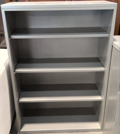 A four-shelf gray metal bookcase or cabinet shown in a furniture store setting, with wood cabinets in the background and a light-colored upholstered chair partially visible on the left.