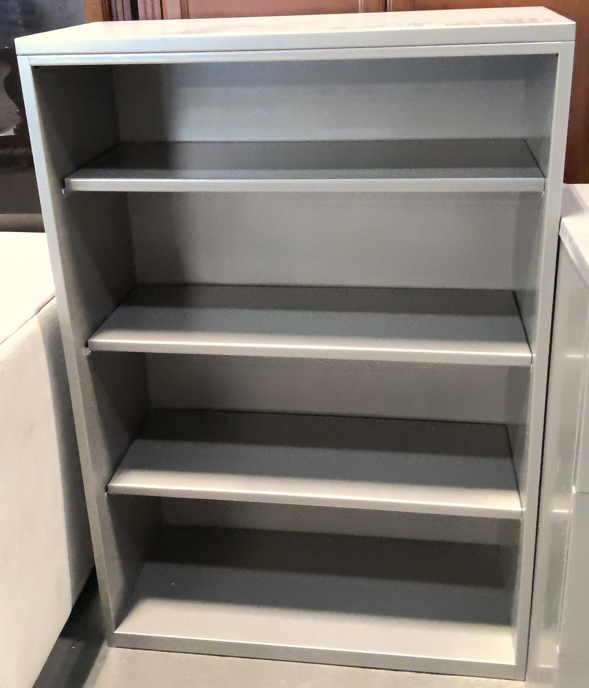 A gray metal 4-shelf bookcase standing in a furniture store, with wooden cabinets in the background.