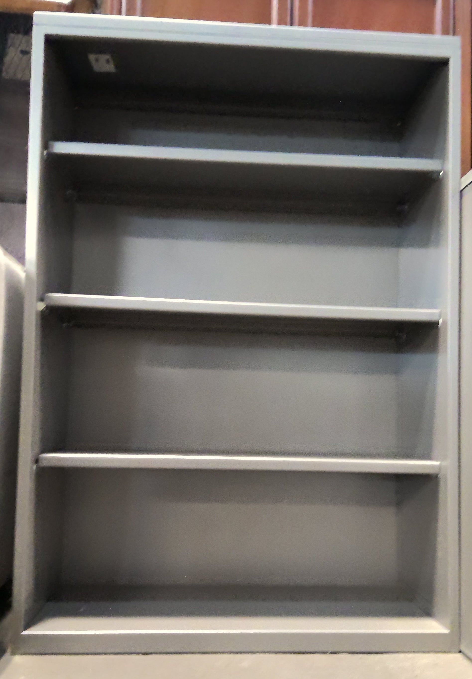 A tall gray metal storage cabinet with four shelves, shown standing in what appears to be a warehouse or storage room with wooden cabinets in the background.