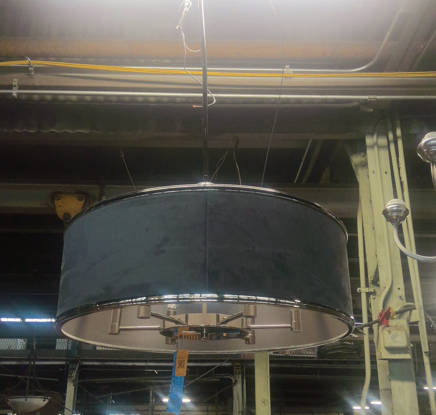A large drum-shaped black fabric pendant light shade hanging in a cluttered warehouse or showroom environment with bright overhead lights and industrial ceiling details.