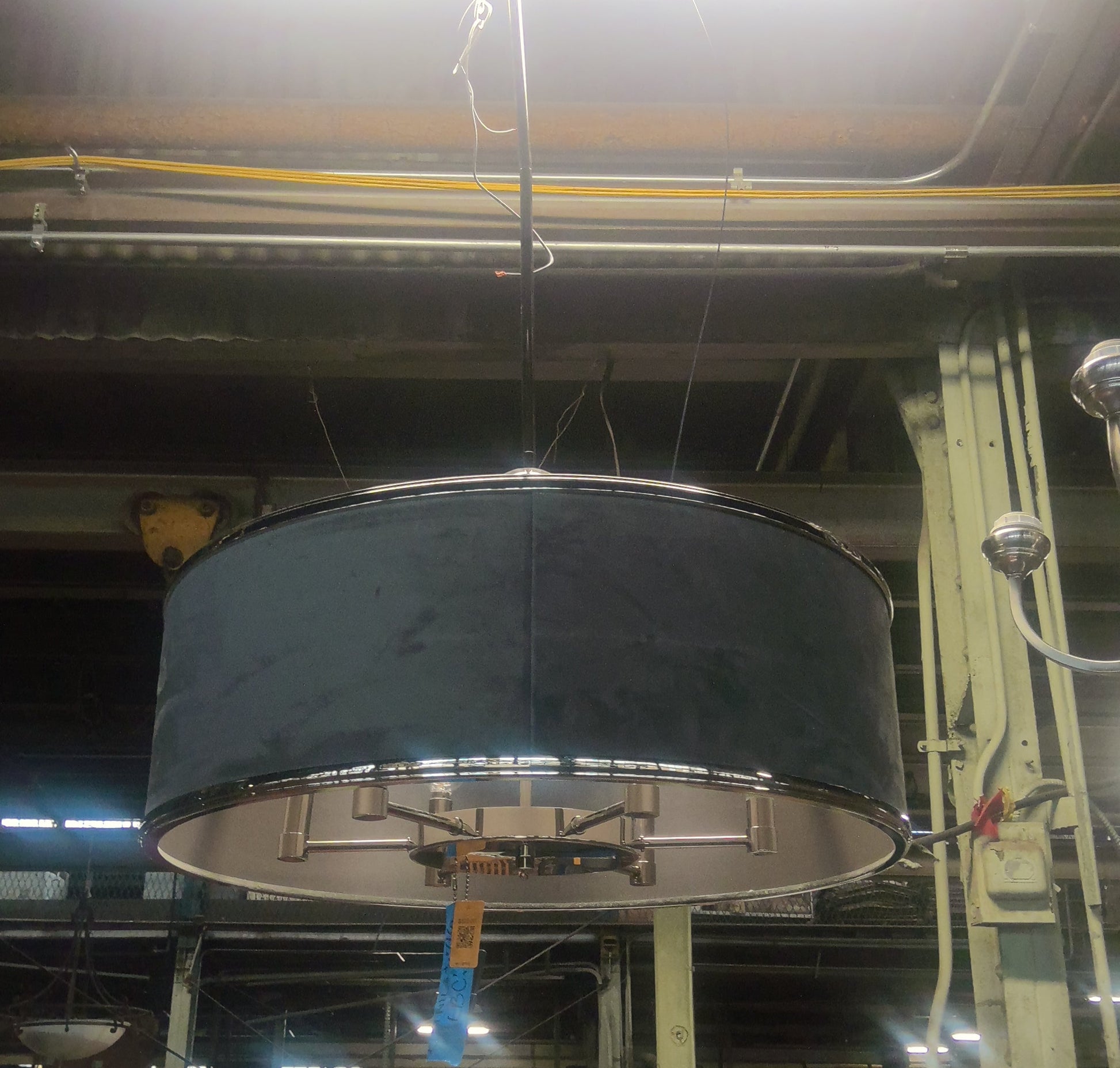 A large drum-shaped black fabric pendant light shade hanging in a cluttered warehouse or showroom environment with bright overhead lights and industrial ceiling details.